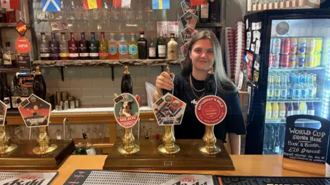 A woman with long brown hair stands behind a bar with her hand resting on a beer tap. 