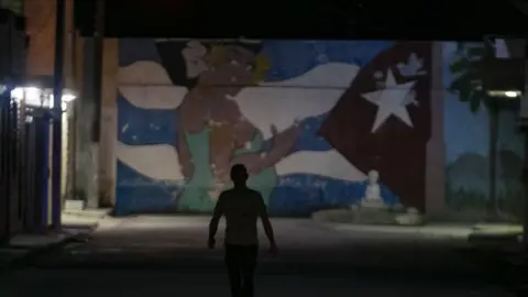 Getty Images A pedestrian walks down the street during a rolling blackout in Havana. He is in the dark but the mural stands out in front of him. The mural features a woman wearing a green dress and a Cuban flag motif.