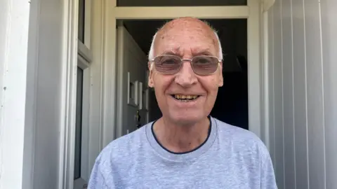 Nicola Haseler/BBC A man in his 80s wearing glasses and a grey T-shirt stands at his front door. He is looking directly at the camera and smiling.