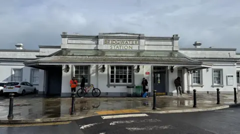 Picture looking front on at Bridgwater Station. It's the front of the station and an old-white building which is a Victorian building. There's some people in the distance waiting around outside the building waiting for trains or to be picked up.