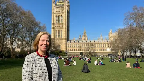 Ruth Cadbury MP berdiri di sebuah taman dengan Westminster di latar belakang.