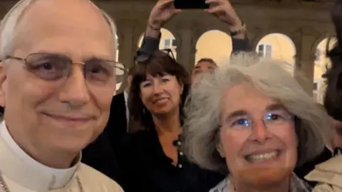 Pope Leo XIV, Professor Anna Rowlands and Sister Nathalie Becquart smile for a selfie inside the Vatican. The Pope is wearing white attire and has glasses. Anna Rowlands is dressed in a black shirt and has a straight brown hair with a fringe. Sister Nathalie Becquart has short grey hair and thin-rimmed glasses. She is wearing a grey shirt. 
