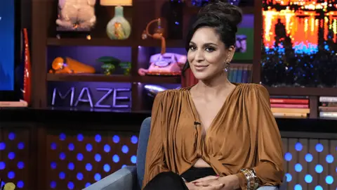 Getty Images Jessel Tank, a woman wearing a brown top, sitting on a blue softa, looking at the camera with a smile. Behind her is a sparkly background, with a letter arrangement spelling out "maze", books, a teddy bear and a landline phone.