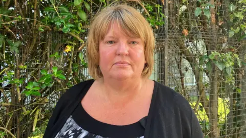 Niree McMorris has long blonde hair. She is standing in front of a fence. She is wearing a black cardigan and grey top.