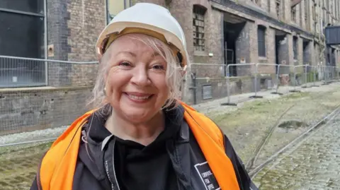 Lynn Saunders has blonde hair and is wearing a black jumper, a black jacket and a high visibility orange vest and white hard hat.