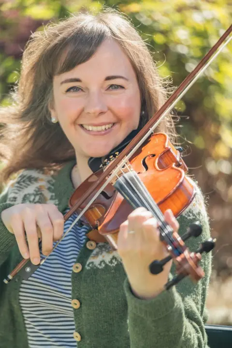 Mark Craig Claire White looks at the camera while playing a violin on a sunny day.