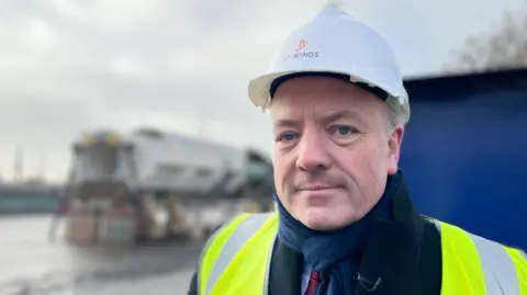 A man wearing a suit, a hard hat and a yellow hi-vis vest stands on a dockside