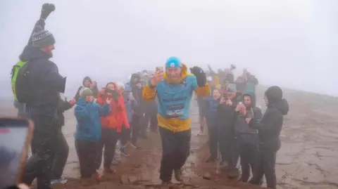 Chris Daniel Chris Daniel wearing a blue t-shirt and under a yellow long sleeved shirt with a blue hat. Two rows of people line the pathway either side of him cheering him on and some are filming him at the summit of Pen y Fan. There is fog behind them obstructing the view of the background.
