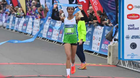 Pacemaker Selsouli has the number 26 pinned on her top. She's holding up a blue ribbon as she runs through a finishing line. Crowds are watching from the sidelines. 