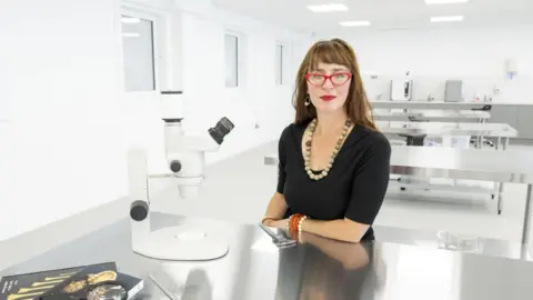 ODAIT A woman with red glasses a black top and a low hanging stone necklace puts her left hand on a metal table. On the table is a white microscope. She has long red hair and wears blue, orange and white. 