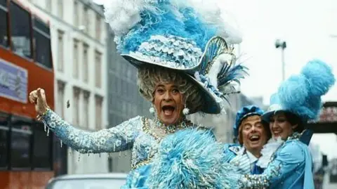 Stanley Baxter stands at a bus stop, dressed as a panto dame in a blue dress and large hat. He is sticking his leg out into the road comically, as a large red bus with a Strathclyde Transport sign drives by. Two other cast members stand behind him, laughing.