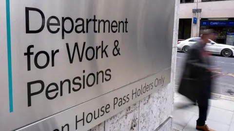 An out-of-focus person walks down a street with the sign for the Department for Work and Pensions in the foreground, outside Caxton House in London on 19 February