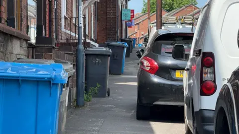 Bins left out on Stepping Lane in Derby, obstructing the pavement alongside cars parked on the pavement.