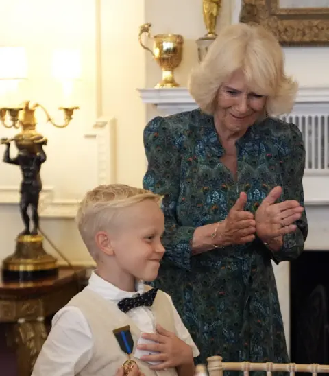 PA Media Rupert Williams standing in front of Queen Camilla. Rupert wears a white short sleeved shirt, a beige waistcoat and a black bow tie. Queen Camilla wearing a patterned green, blue and orange dress. Rupert smiles as his medal is pinned to his shirt. Camilla appears to be clapping. 