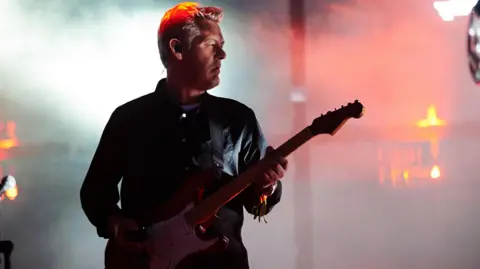 Angelo Bruschini playing guitar at Glastonbury Festival in 2014. He is looking away from the camera and smoke from the stage is behind him.