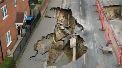 An aerial picture of a large sinkhole which has opened up beneath one lane of the road. The road surface has collapsed into the hole and a broken pipe and small pool of water are visible. Two sections of the adjoining pavement have also begun to collapse into the hole.