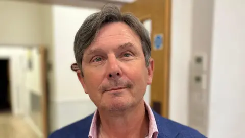 A man called Andy Sloan. He has neatly trimmed dark-greying hair. He is stood in front of a double doorway and is wearing a navy blue jacket and a pink shirt.
