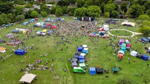 GHRVISION An aerial view of a huge park, with lots of people sitting down and walking around tents and gazebos. Each gazebo is a different colour and towards the back is a large stage with speakers. Also visible are coloured deck chairs, fun fair games and some parked vehicles.