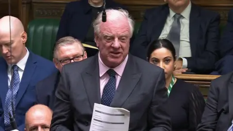 Sir Edward Leigh wearing a dark grey blazer and pink shirt with a blue tie that has white dots on. He is standing up holding a piece of paper with several other parliament members sat around him on green benches.