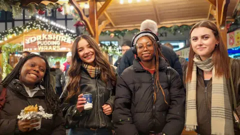 Four young women stand in a row smiling at the camera at a Christmas market stall in Kingston upon Thames, one of them Amelia Shannon, holding a cup of mulled wine. In the background there is a 'Yorkshire pudding wraps' stall visible.