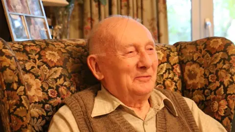 Tom Hall An older man, with a brown top over a light coloured shirt. He is sat in a large floral chair and is smiling