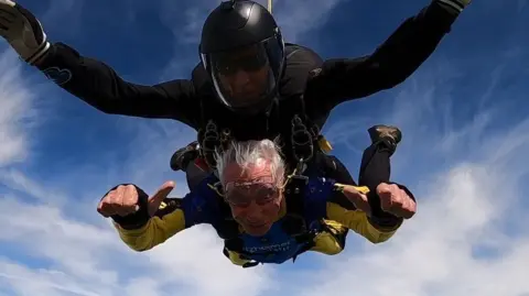Brain, who has grey hair and wears and yellow and blue boiler suit, gives two thumbs up to the camera as he skydives. He is attached in tandem to an instructor, who is wearing all black. 