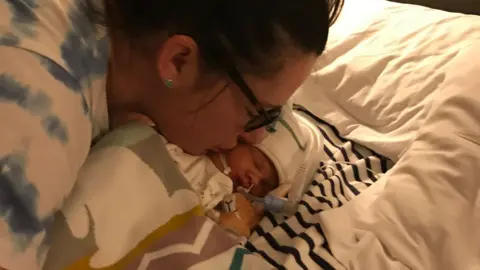 Sarah Robinson, who is wearing glasses and has dark hair tied back in a ponytail, kiss the cheek of newborn Ida Lock, in a hospital bed.