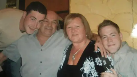 Supplied Two young men with a man and woman who are older. They all have their arms around each other and are smiling at the camera.