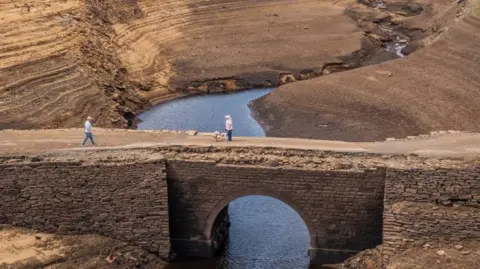 Two people on a stone bridge set amongst the steep sides of a semi-empty reservoir. The bridge is normally submerged but falling levels have made it passable.