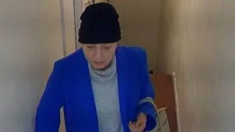 Bedfordshire Police CCTV image of a woman, in a hallway, wearing a grey jumper, blue coat, with a dark-coloured hat on her head. She has a bag over her left arm. She is looking ahead. The walls are pale around her. 