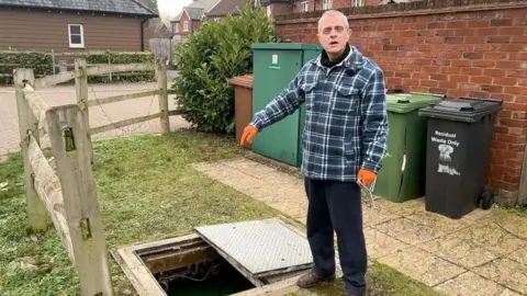 Stuart Tyrrell is wearing a blue checked overshirt and orange work gloves. He is pointing into a hole created by the removal of a manhole cover. It is a residential area and behind him are brown, green and black wheelie bins.