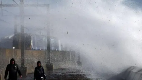 Getty Images Two men, wearing black jackets with their hoods up being covered with water from a large white wave.