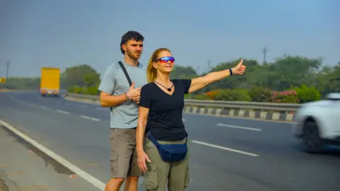 BBC/Studio Lambert Tom and Caroline are stood hitchhiking on the side of a road
