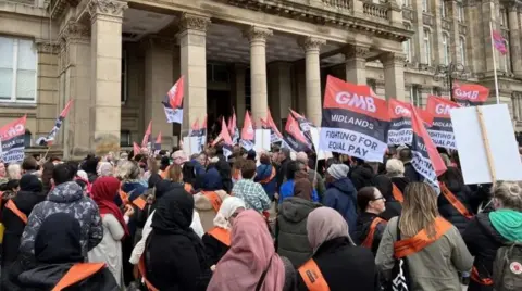 Protestors gathered outside Birmingham Council House. Many are wearing orange sashes and some are carrying flags which have the GMB union logo on them as well as text which reads 'fighting for equal pay' in black lettering.
