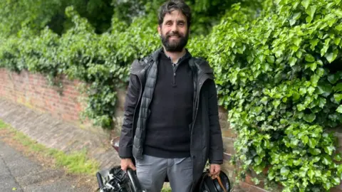 A portrait image of Miguel stood on a pavement by the QMC roundabout. He is holding a folded bicycle in one hand and a helmet in the other.