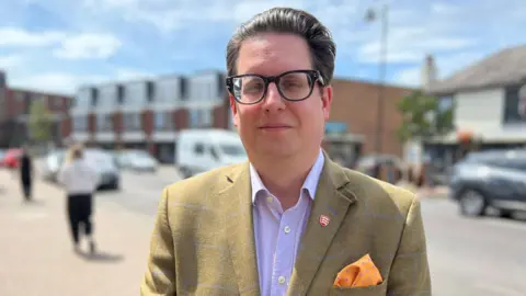 Stuart Woodward/BBC A man wearing a checked light brown suit jacket and a purple shirt. He has black-framed glasses. He looks at the camera. A road is behind him.