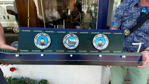 Whitchurch Waterway Trust A wooden replica of a canal boat held up by two people outside the window of a building. It is dark green and blue and has images of three ceremonial plates on the side.