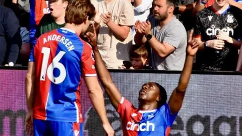 HY MONEY/ TOPFOTO Une photo d'un joueur de football de Crystal Palace agenouillé sur le terrain pour célébrer, les bras levés, tandis qu'un autre coéquipier se tient à proximité. Les deux joueurs portent la tenue rouge et bleue de Crystal Palace. En arrière-plan, les spectateurs applaudissent dans les tribunes.