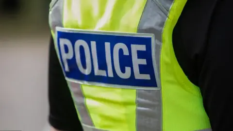 The back view of a police officer wearing a yellow vest with the words police in bold white lettering against a blue rectangular background.