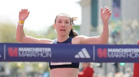 Manchester Marathon Naomi Robinson raises her arms up as she looks at the finish tape, which has the words Manchester Marathon across it. She wears a running top and has her hair tied back in a ponytail.