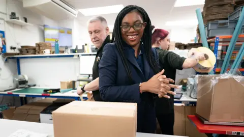 Kemi Badenoch in a factory in Brentwood with two employees behind her as they box up goods.