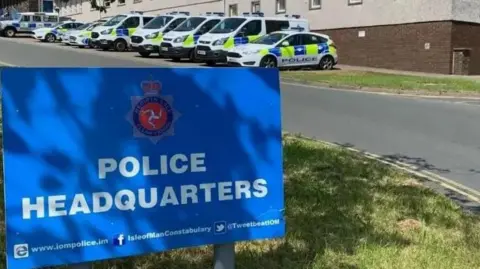 In the foreground you can see a large blue sign which reads police headquarters. In the background, about 9 police cars and vans park up in a row. 