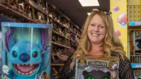 A woman holding a toy and smiling. Another toy is prominent on the photo to her left. Toys are on shelves in the background.