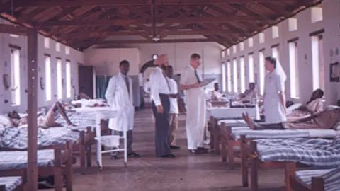 Éanna Mac Cana/Little Ease Films A old, colour wide shot photo of Dr Denis Burkitt looking at a chart in a hospital ward in Africa. He is wearing a white collared shirt, a dark tie and white trousers. There is a nurse in a white uniform standing across from him. Behind Dr Burkitt are three other doctors. The roof has bare wooden beams, and there are some patients wearing gowns in the beds.