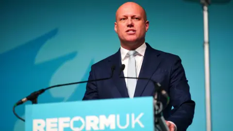 PA Dan Thomas speaking at a podium which says "Reform UK". He is wearing a dark blue suit and light blue tie, over a white shirt, and is in front of a light teal backdrop.