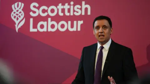 PA Media Scottish Labour leader Anas Sarwar standing in front of a red wall with the party name in white. He is wearing a dark-coloured suit and is looking right at the camera.