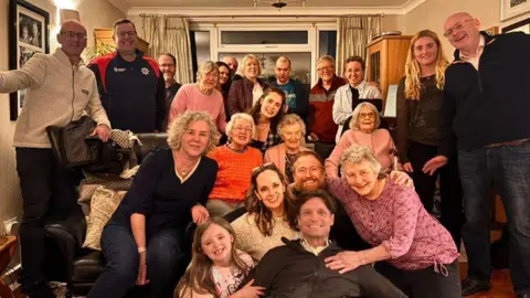 Hans Obma 22 people stand in frame in a living room. Hans Obma is at the centre right at the bottom. Everyone smiles at the camera. 