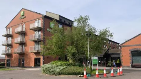 LDRS On the left is a four-storey red brick building with glass balconies. On the building is a green Holiday Inn logo and signage. In the middle is a landscaped lawn which is half surrounded by red and orange cones.