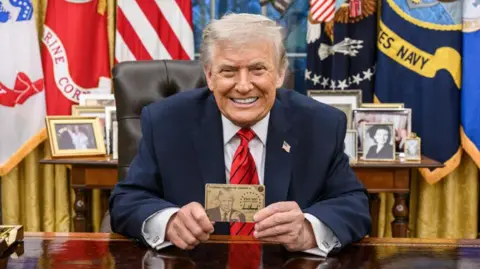 @realDonaldTrump/Truth Social President Donald Trump smiles for the camera in the White House and he holds up a Trump Gold Card 