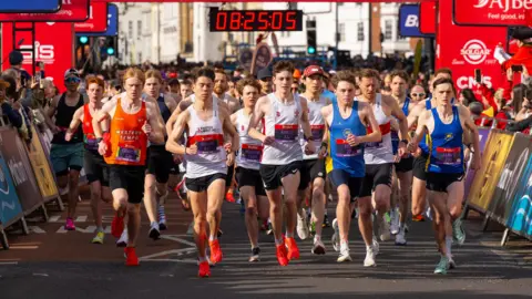 AJ Bell Great Bristol Run Dozens of people running down a road in Bristol. There is red canopy with the AJ Bell Great Bristol Run logo which has the race time on the clock.